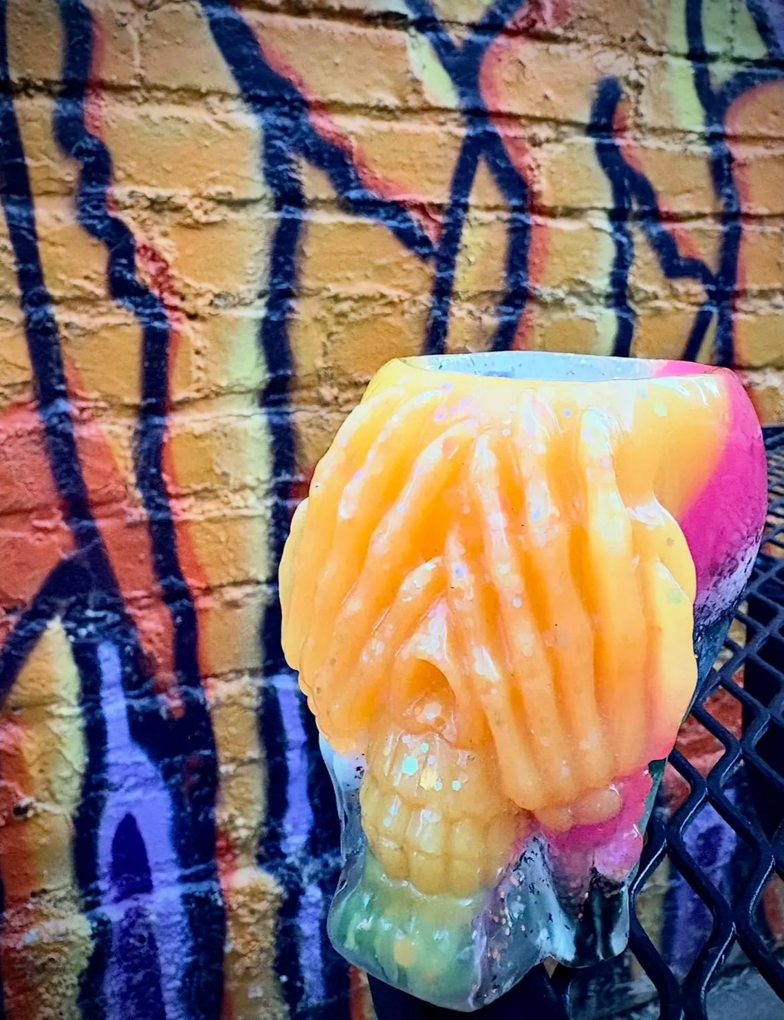 Orange and pink resin skull cup with sculpted hands over its eyes, in front of a graffiti wall